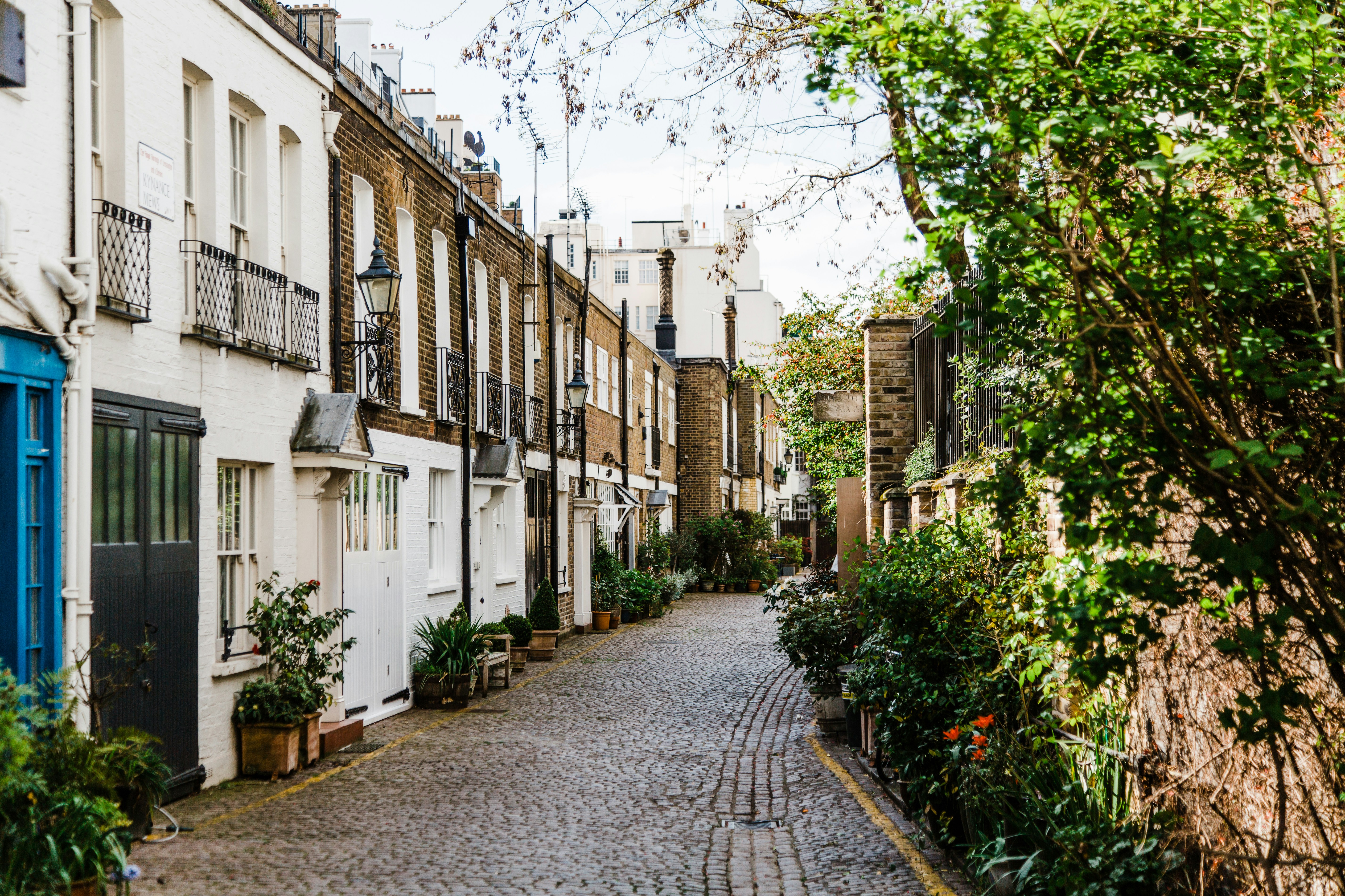 Charming cobblestone street lined with traditional townhouses and potted plants in a quiet, leafy residential area. The buildings feature brick and white facades with wrought iron balconies, creating a cozy and picturesque urban scene.