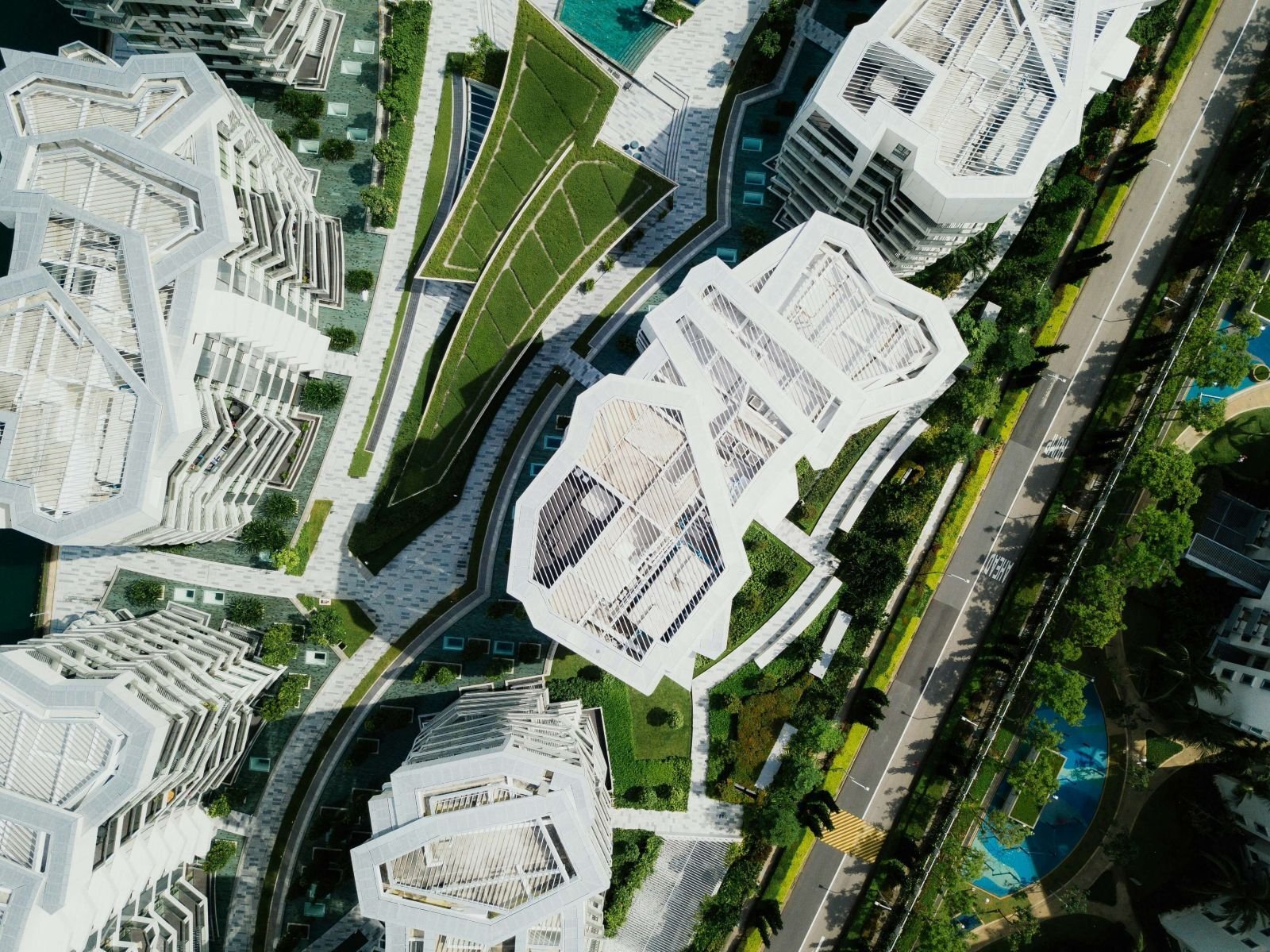 Top-down aerial view of modern high-rise buildings with geometric white rooftops and green rooftop gardens. The layout includes landscaped pathways, roadways, and tree-lined streets, showcasing a sustainable urban design with integrated green spaces.