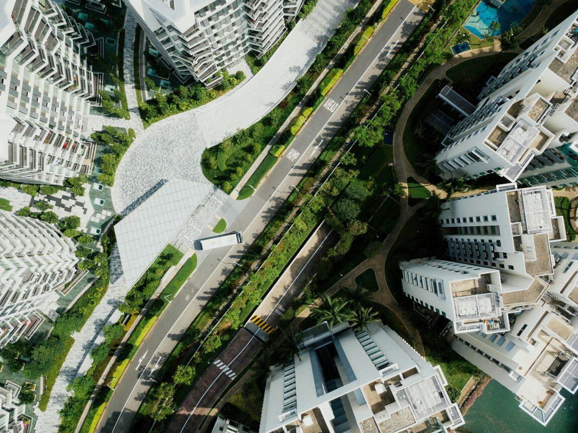Aerial view of a modern urban residential area with high-rise buildings, tree-lined streets, landscaped walkways, and green spaces. The layout shows a well-planned blend of architecture and nature, with a bus and crosswalk visible below.