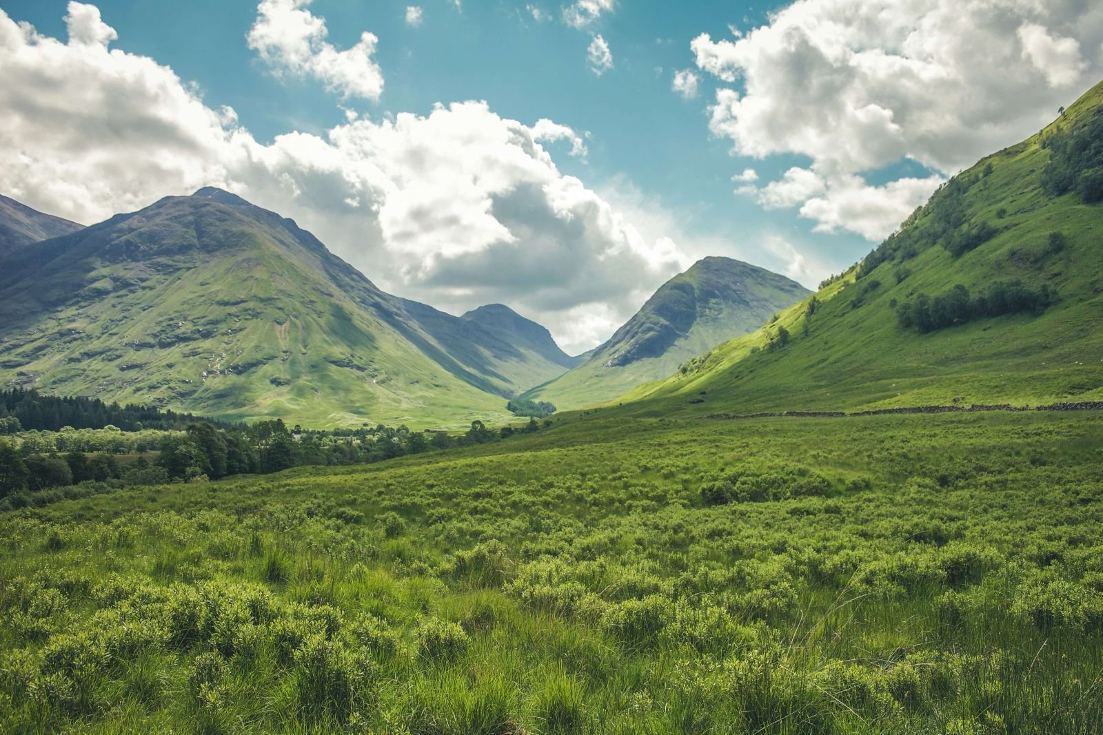 Scenic view of a lush green valley surrounded by steep, grassy mountains under a partly cloudy sky. Sunlight filters through the clouds, casting soft shadows across the landscape, creating a peaceful and expansive highland scene.