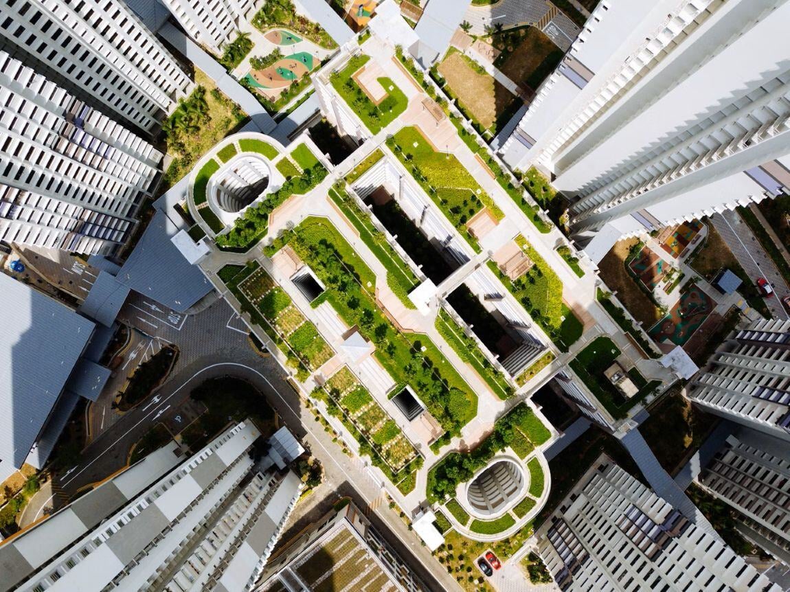 Aerial view of high-rise buildings with green rooftops and landscaped terraces. The central complex features neatly arranged garden areas, pathways, and plants, blending urban architecture with sustainable green space in a dense city environment.