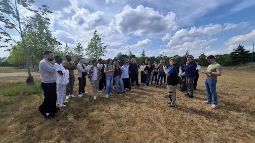 A group of people gathers outdoors on a dry grassy field under a partly cloudy sky, listening to a guide or instructor. Some individuals take notes while others observe. Trees line the background, suggesting the event is part of an educational or environmental field activity.